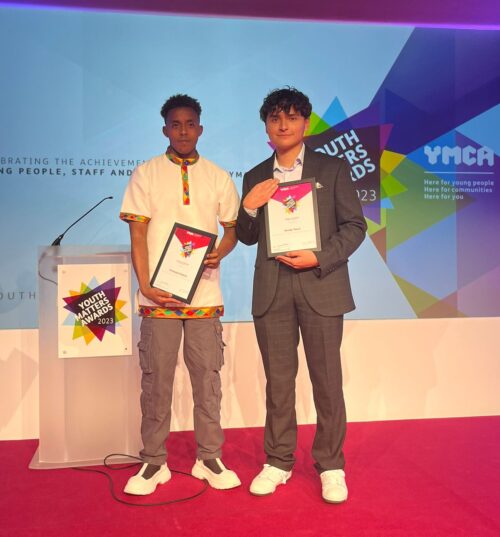At the Youth Matters Awards. Temesgen (left) holding his certificate on stage, wearing a traditional Ethiopian shirt and trousers. Randy (on the right) is holding his certificate smiling, wearing a blue suit.