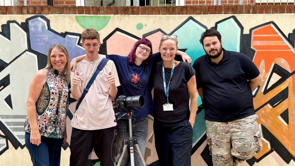 group of 5 people, men and women of various ages, standing in front of a wall with filming equipment