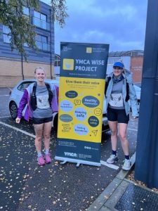 2 women in running attire stood by a banner for What is Sexual Exploitation project (WiSE)