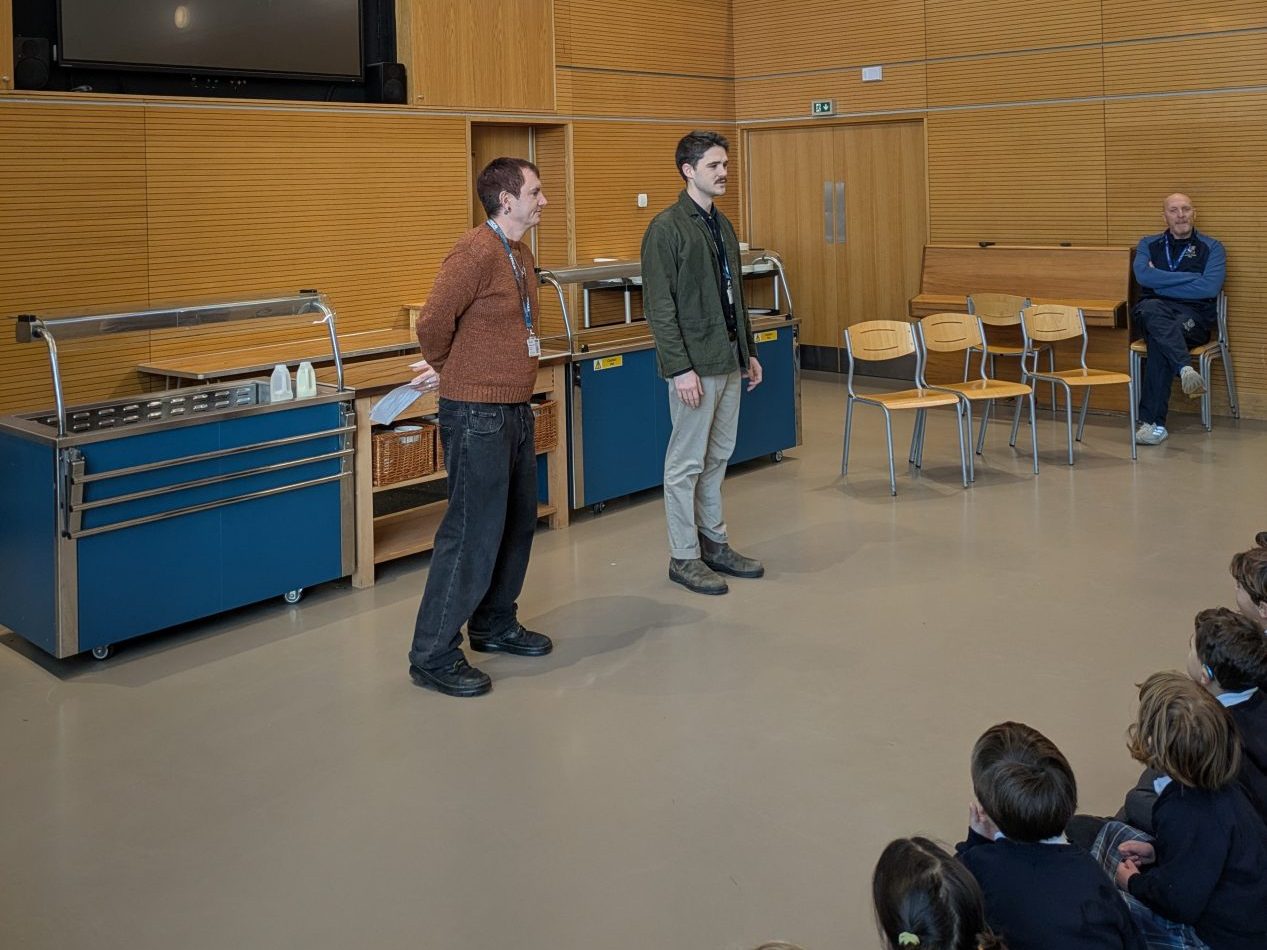 two men standing in front of an assembly of seated children mostly off screen