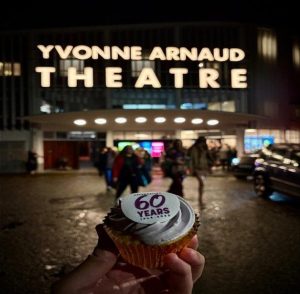 hand holding up a celebration cupcake reading '60 years' in front of Yvonne Arnaud theatre
