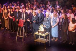 a crowd of people listening to a man stood at their centre near a tiered celebration cake