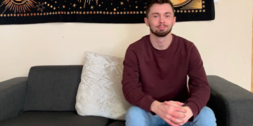 Josh sat on a sofa smiling, wearing a red jumper and blue jeans