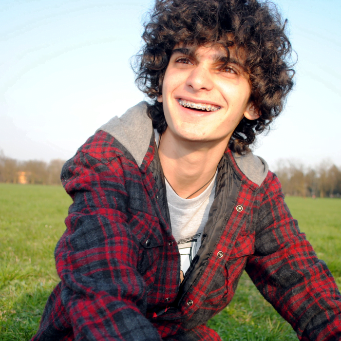 Dylan with curley brown hair and braces smiling at the camera, sat in a grass field