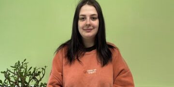 Georgia, smiling stood against a green background with a plant to her side