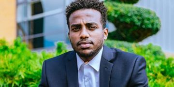 Amanuel, a young man wearing a suit and white shirt with green bushes behind him